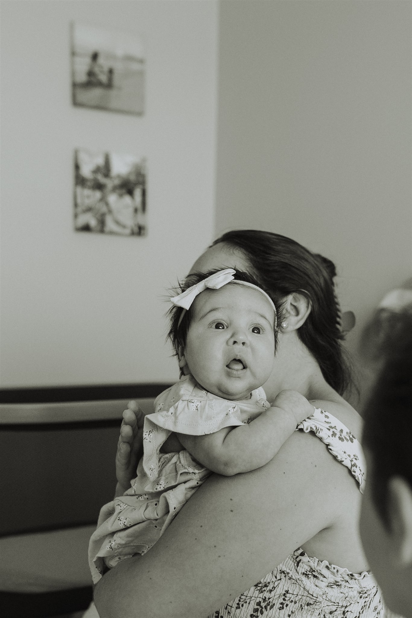 Black and white photo of a baby being held up on moms shoulders with her eyes wide open while her older brother just off camera is looking at her