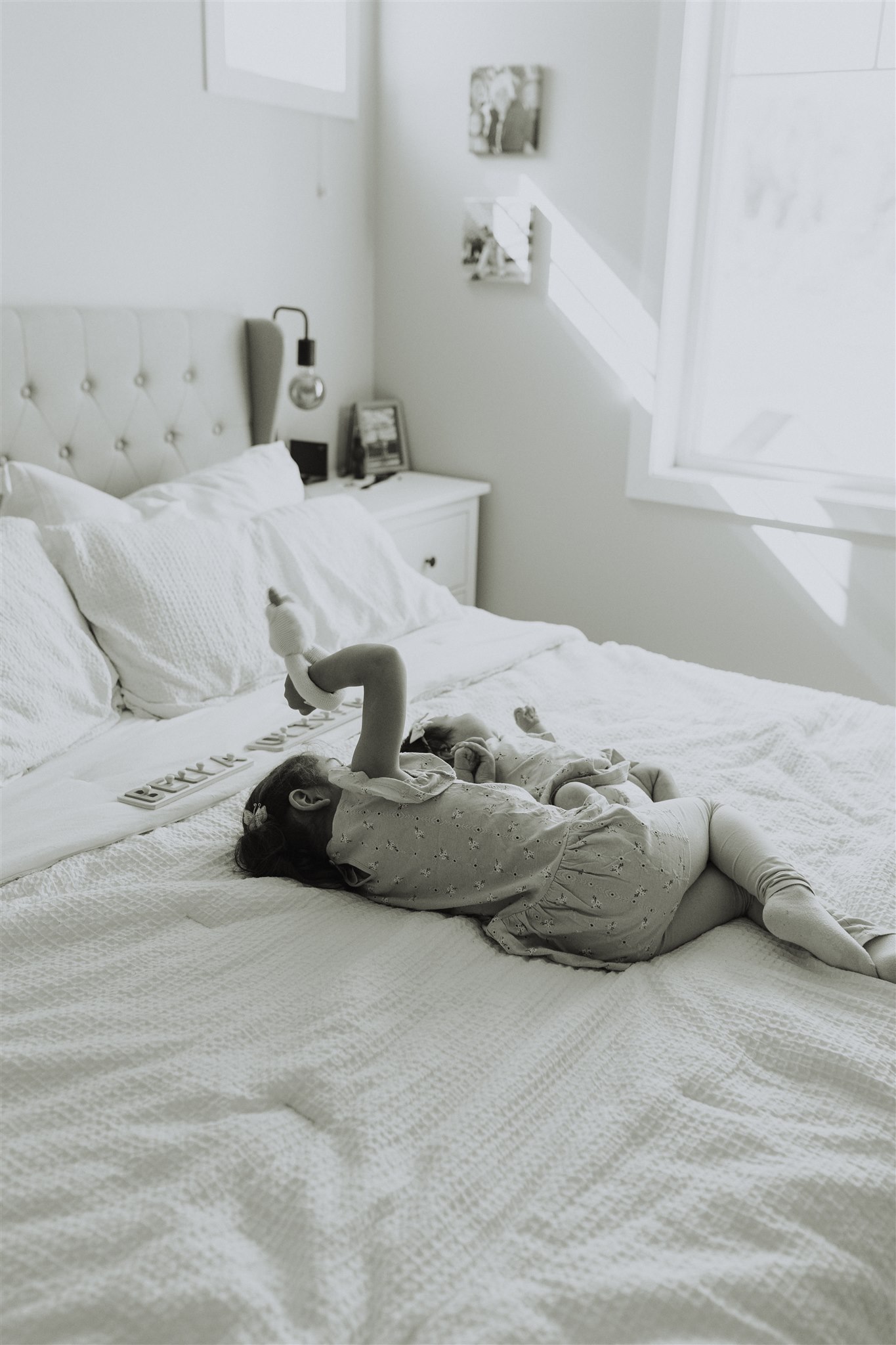 Black and white photo of a big sister and her baby sister laying on the bed while the big sister shows the baby a toy. For a blog about newborn photography with siblings