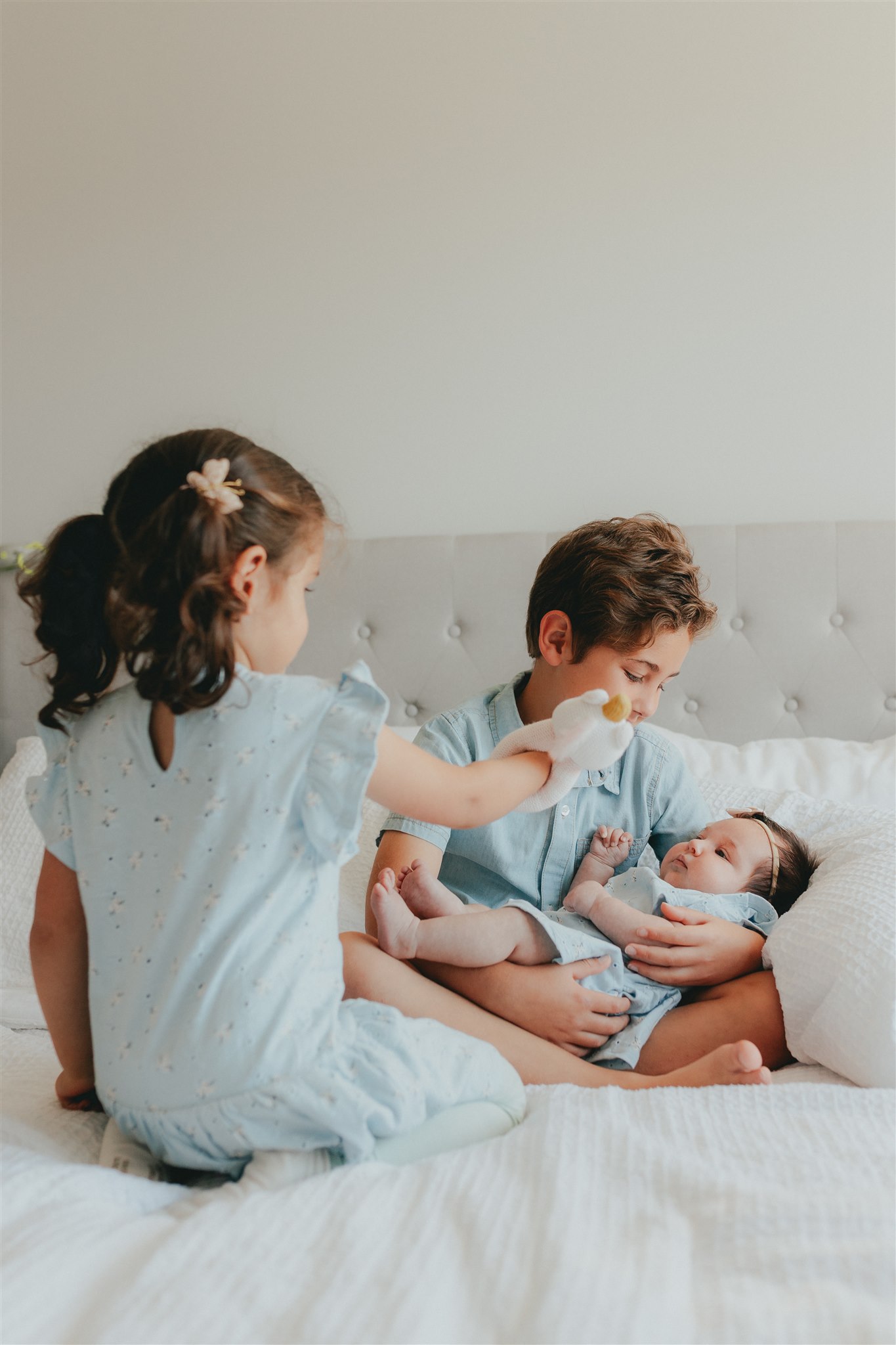 Photo of a boy holding his baby sister and looking at her while the older sister is showing the baby a toy.