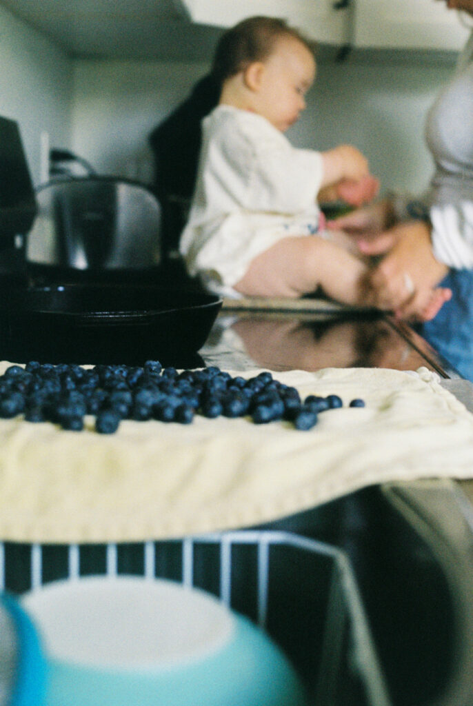 Photo of a baby sitting on the counter with blueberries beside her with the focus of the photo on the blueberries
