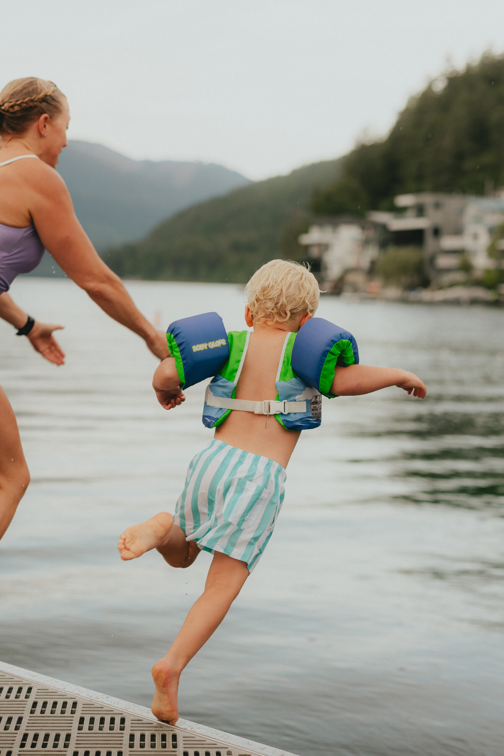 Photo of a mom holding her sons hand while they both jump into a lake off a dock taken from behind