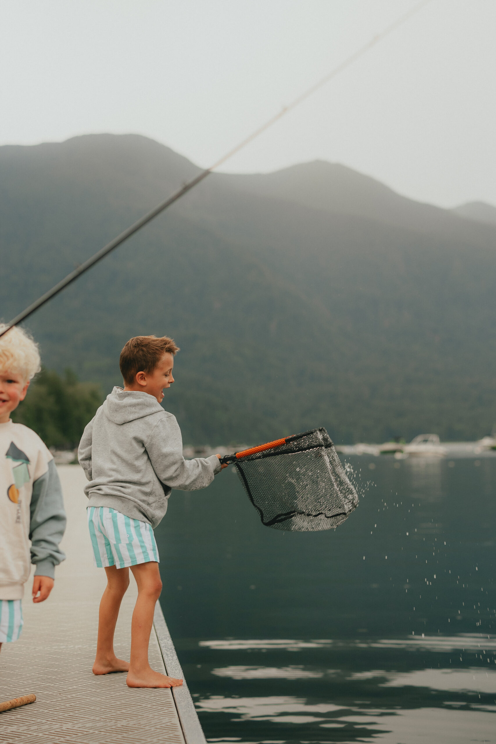 Photo of a little boy standing on a dock holding a fishing net with an excited look on his face while water drips off the net