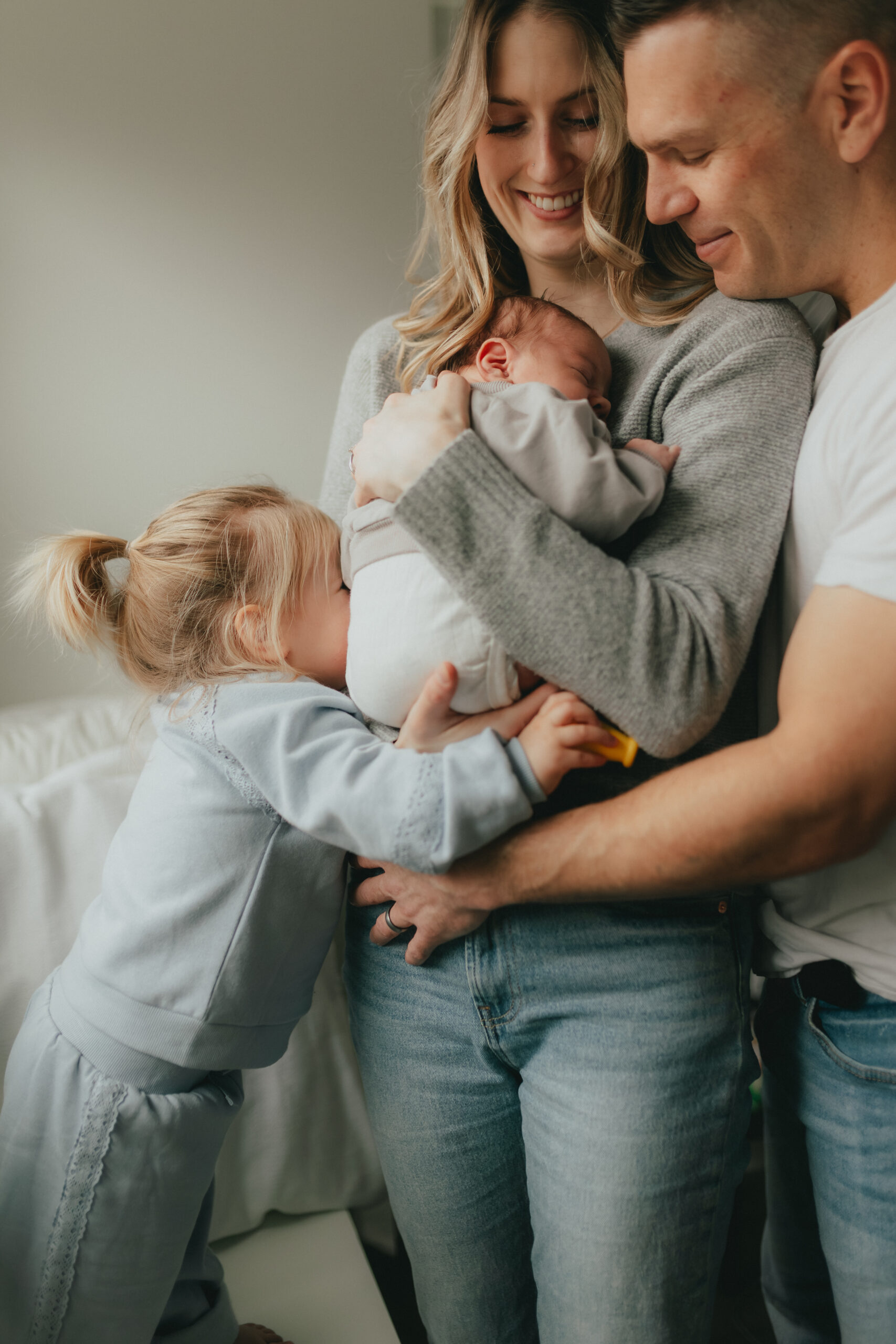 Photo of a mom holding her new baby with dad and her toddler daughter hugging her, with the toddler having her face buried in mom's body. Taken by a newborn photographer, Abbotsford B.C.