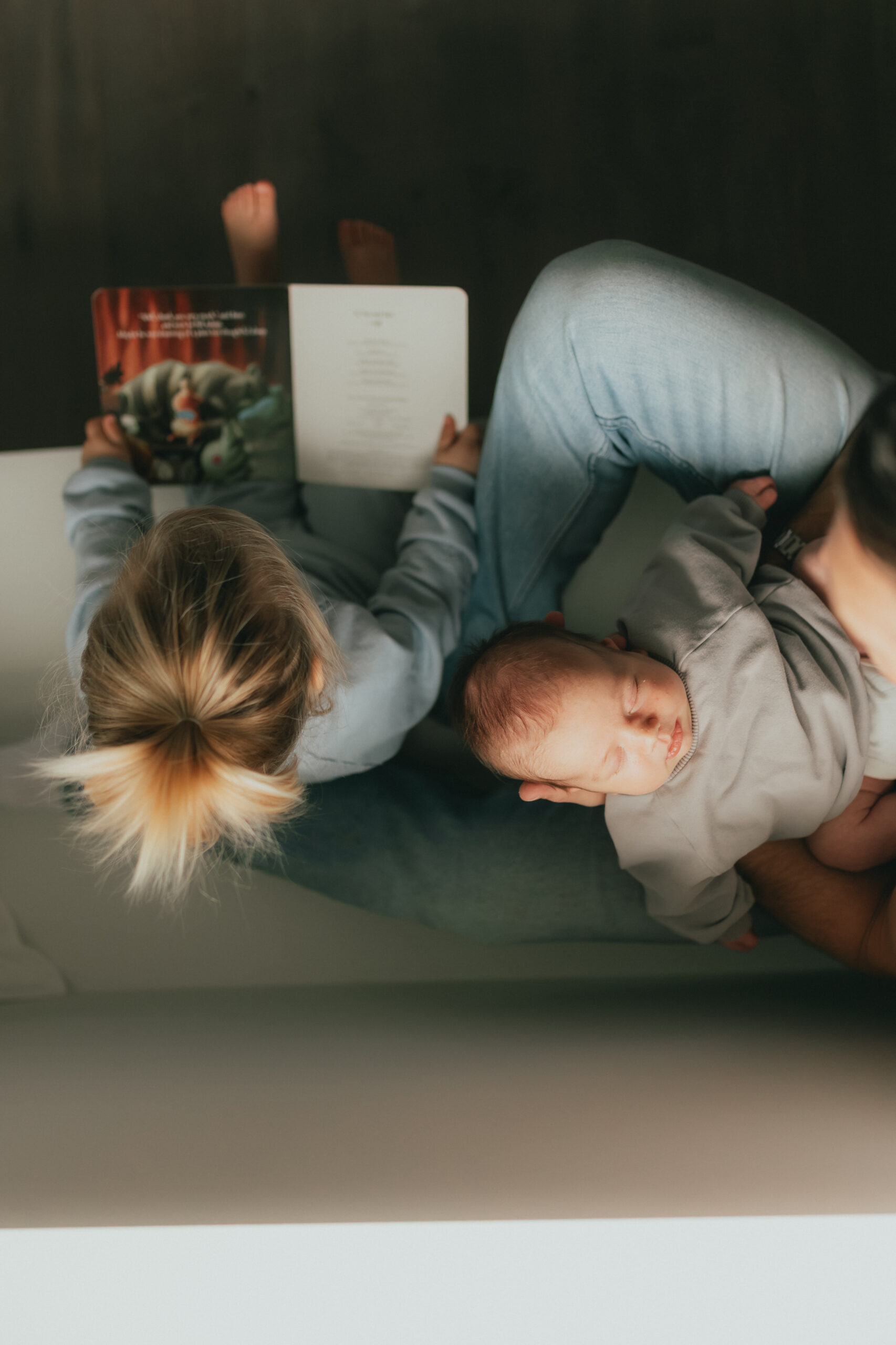 photo of a little girl, her dad and her baby brother sitting on a bench taken from above while the little girl reads a book. Taken by a newborn photographer, Abbotsford B.C.