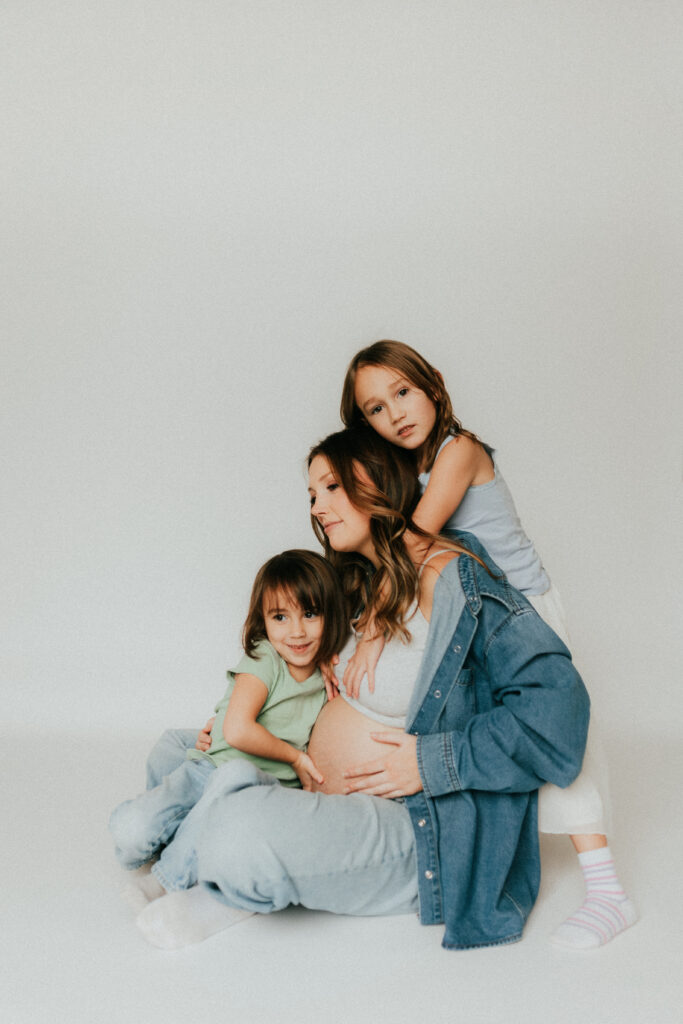 Photo of a mom and 2 daughters sitting on the ground with one girl hugging mom from behind and the other sitting in front hugging moms pregnant belly