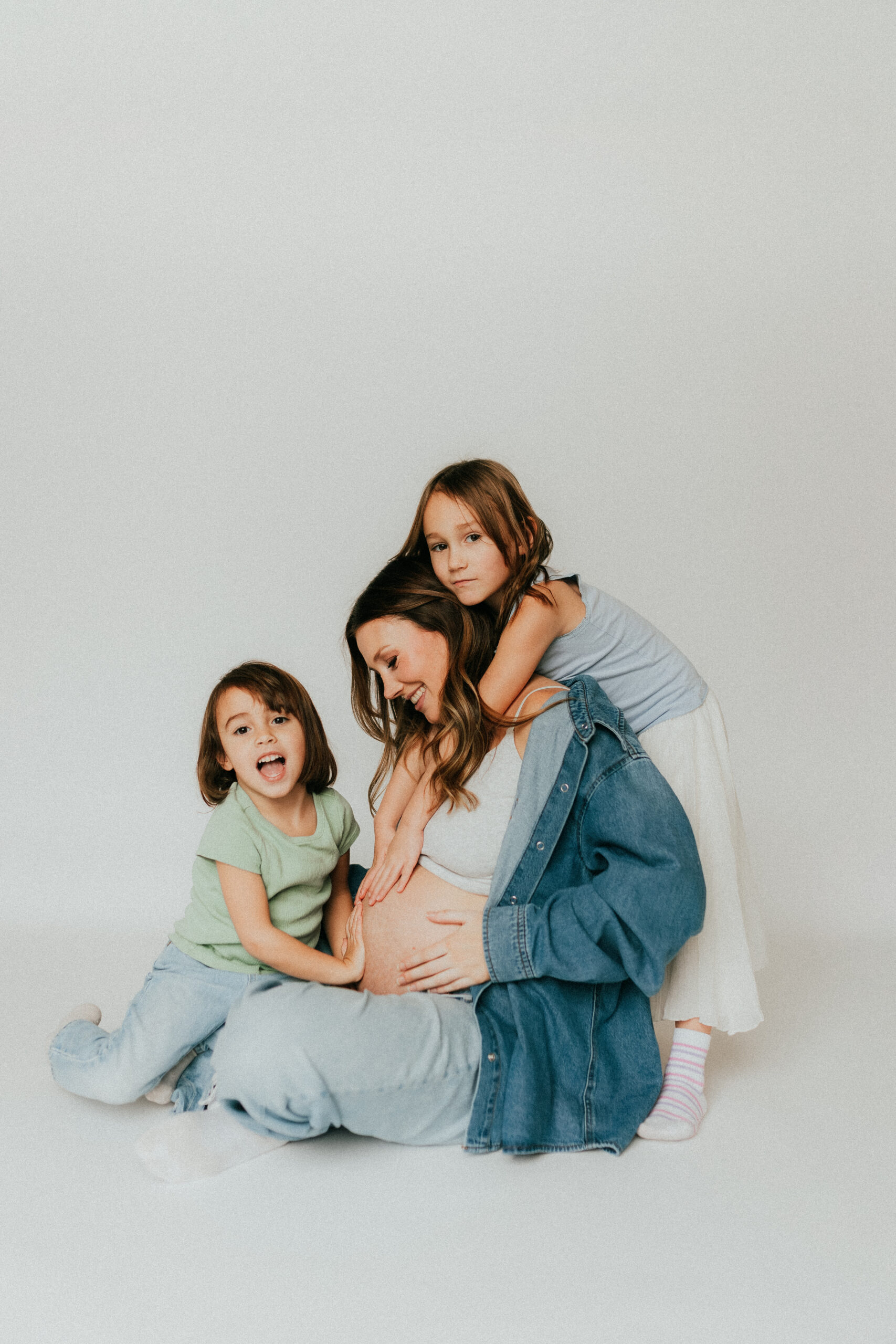 Photo of a mom and 2 daughters sitting on the ground with one girl hugging mom from behind and the other sitting in front hugging moms pregnant belly