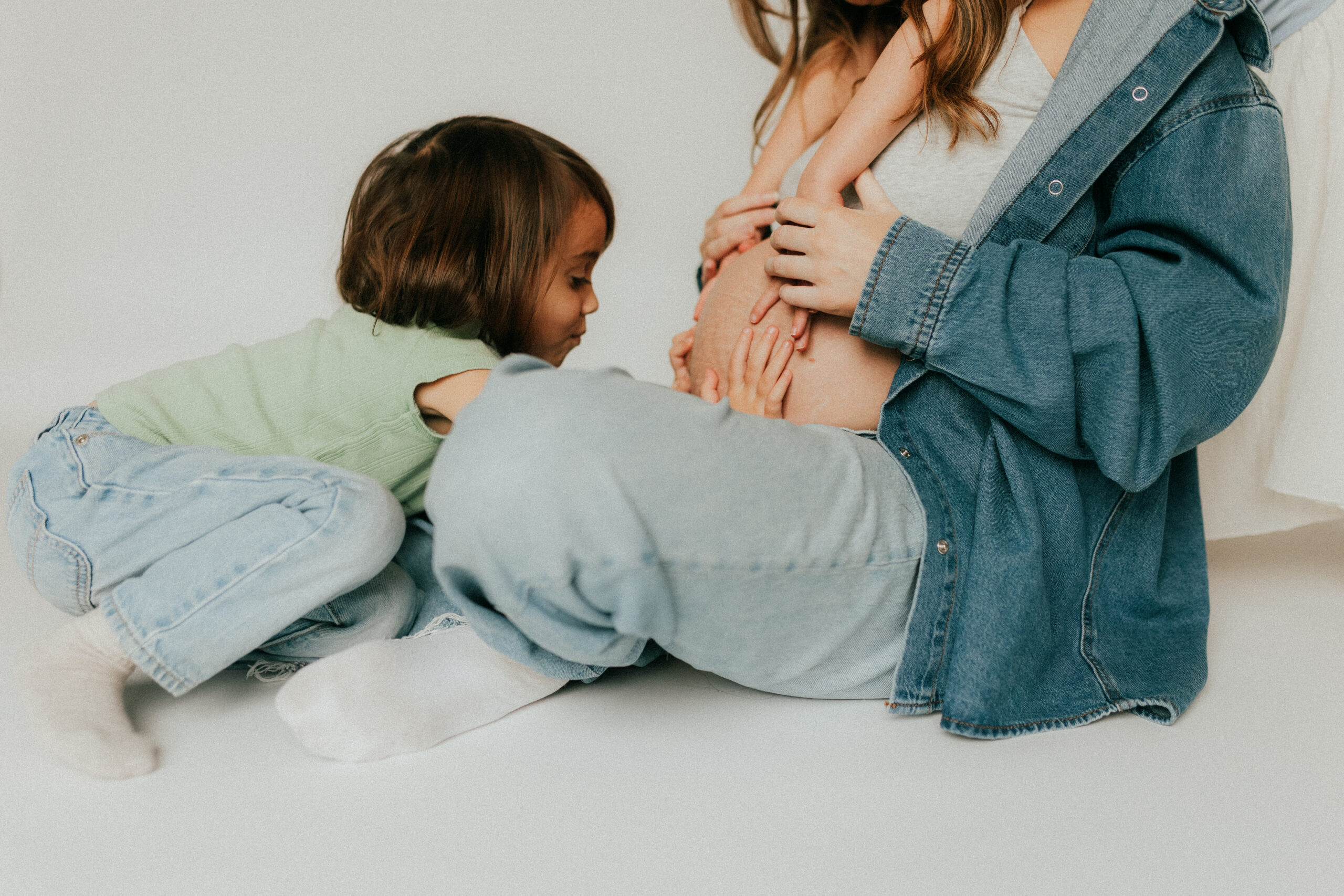 Photo of a mom sitting on the ground with her young daughter sitting in front of her touching her pregnant belly