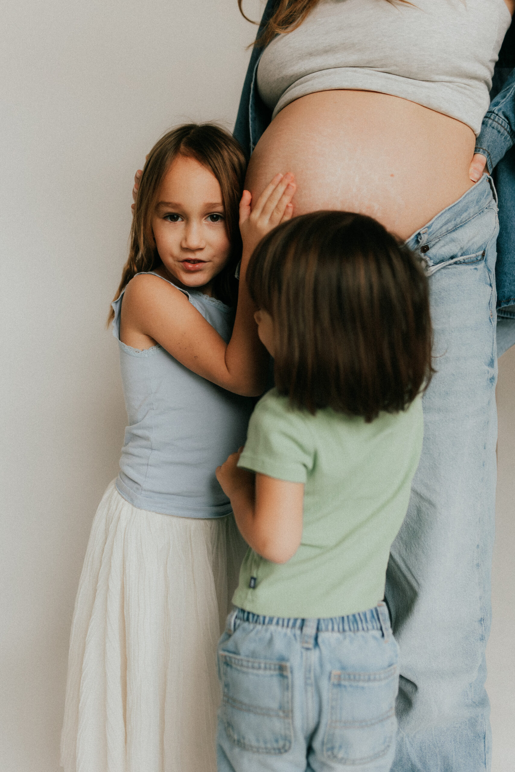 Photo of 2 daughters standing in front of their mom and hugging her pregnant belly
