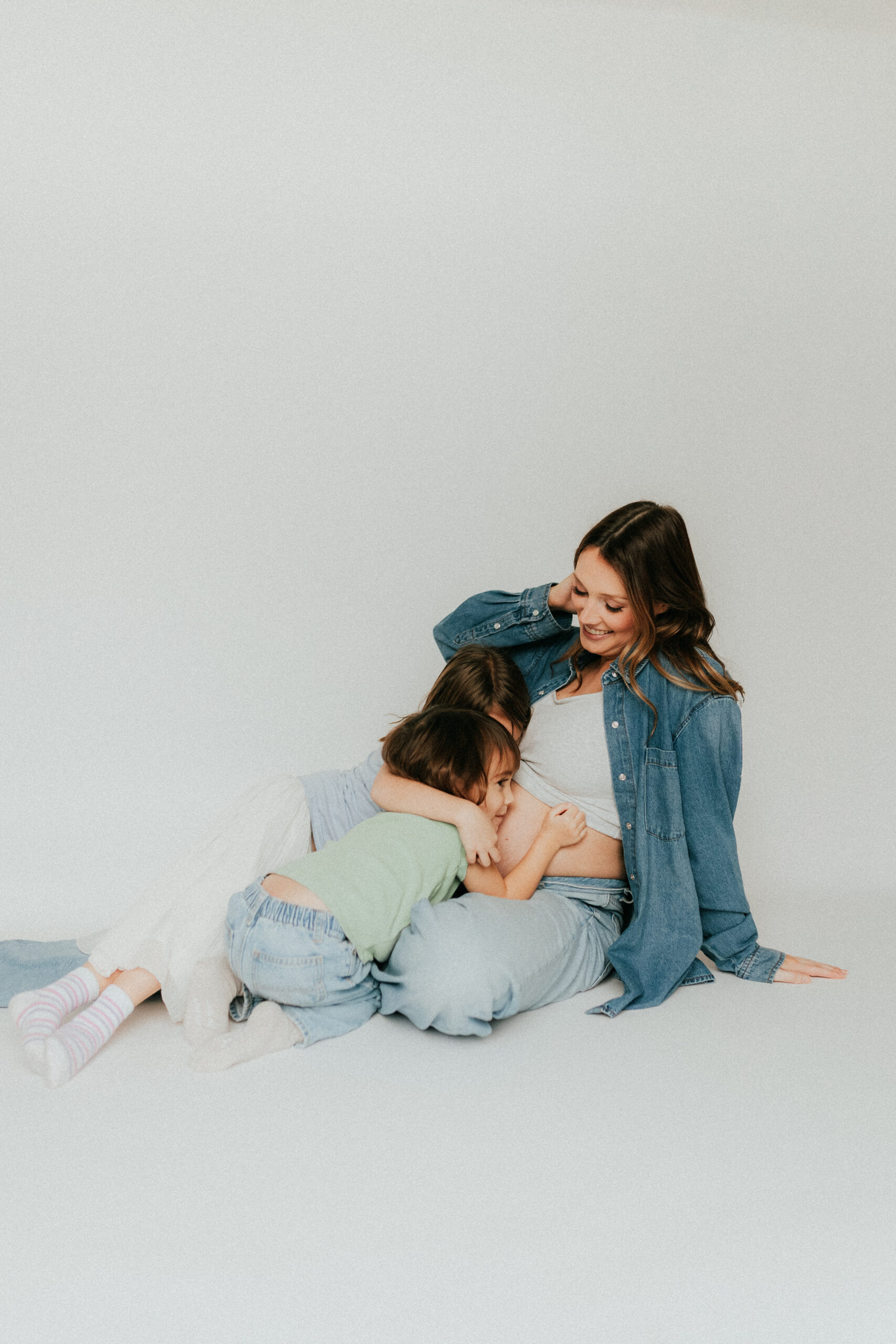 Mom and 2 daughters sitting on the ground while the daughters hug moms pregnant belly
