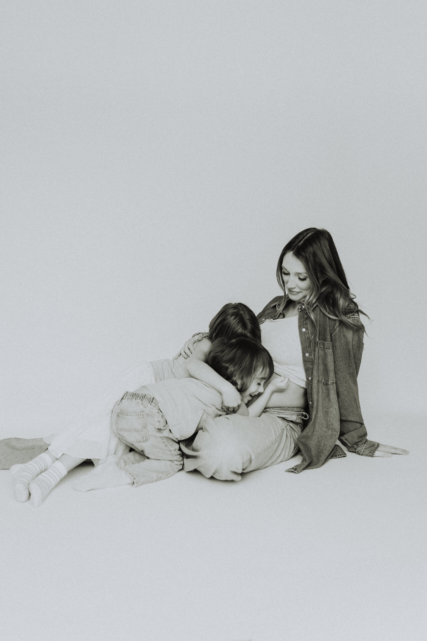 Black and white photo o a mom sitting on the ground with her two daughters hugging her pregnant belly taken at a lifestyle motherhood session in langley