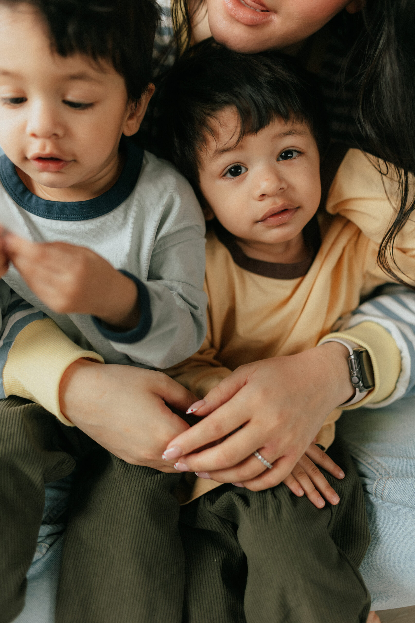 Close up photo of twin toddler boys sitting on moms lap looking at the camera.