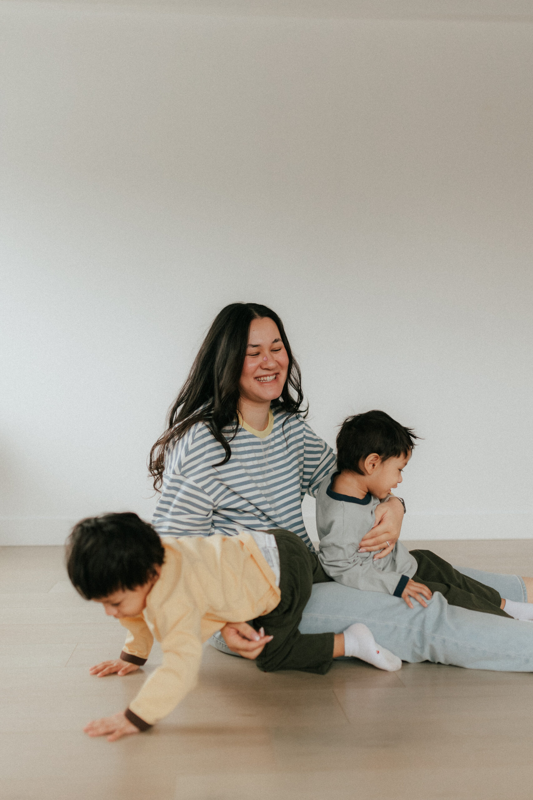 Photo of a mom trying to hold her twin boys while they crawl away and mom is laughing during a motherhood session in abbotsford