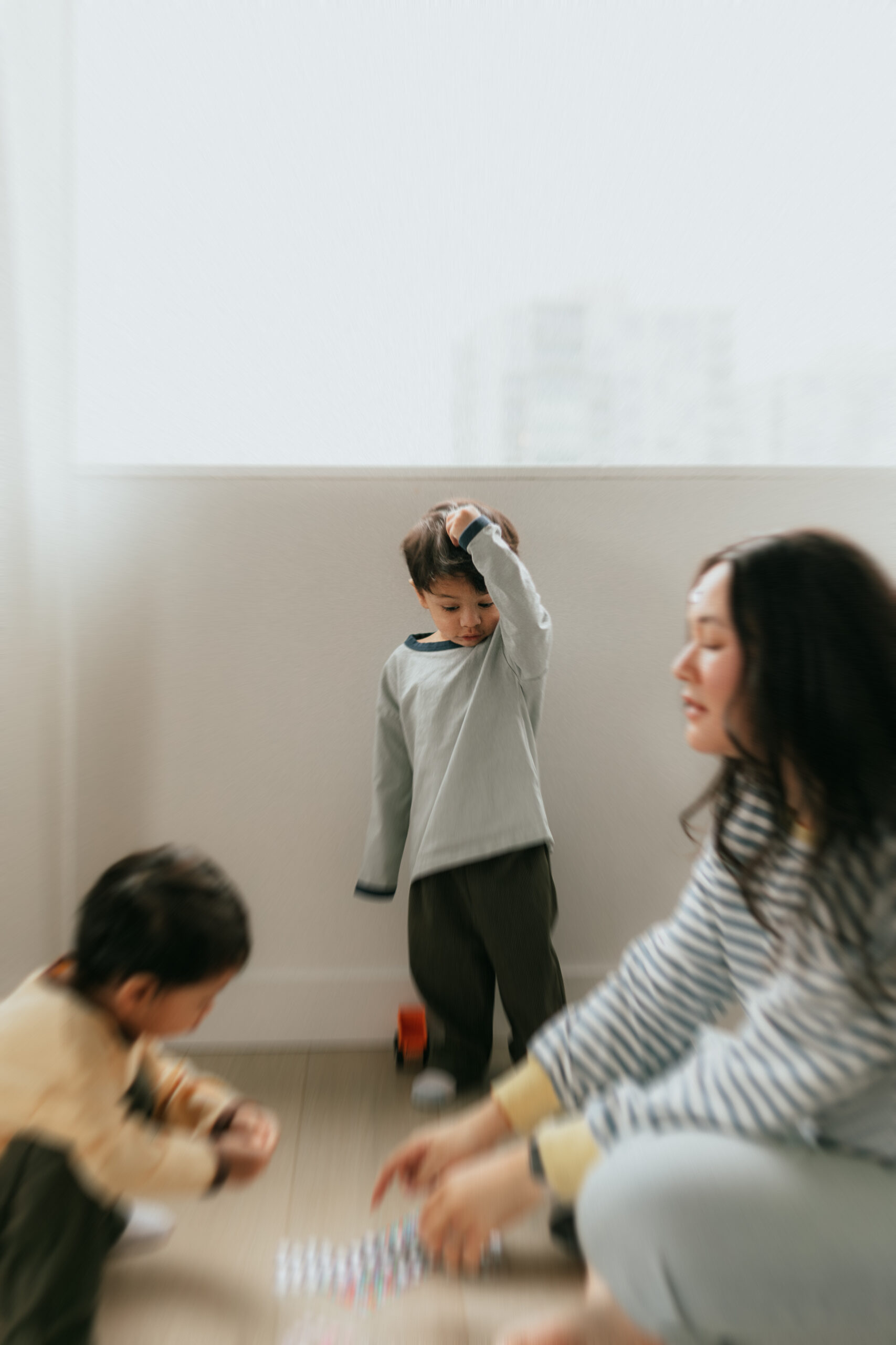 Photo of a young boy holding a toy on his head while his brother and mom play in the foreground.