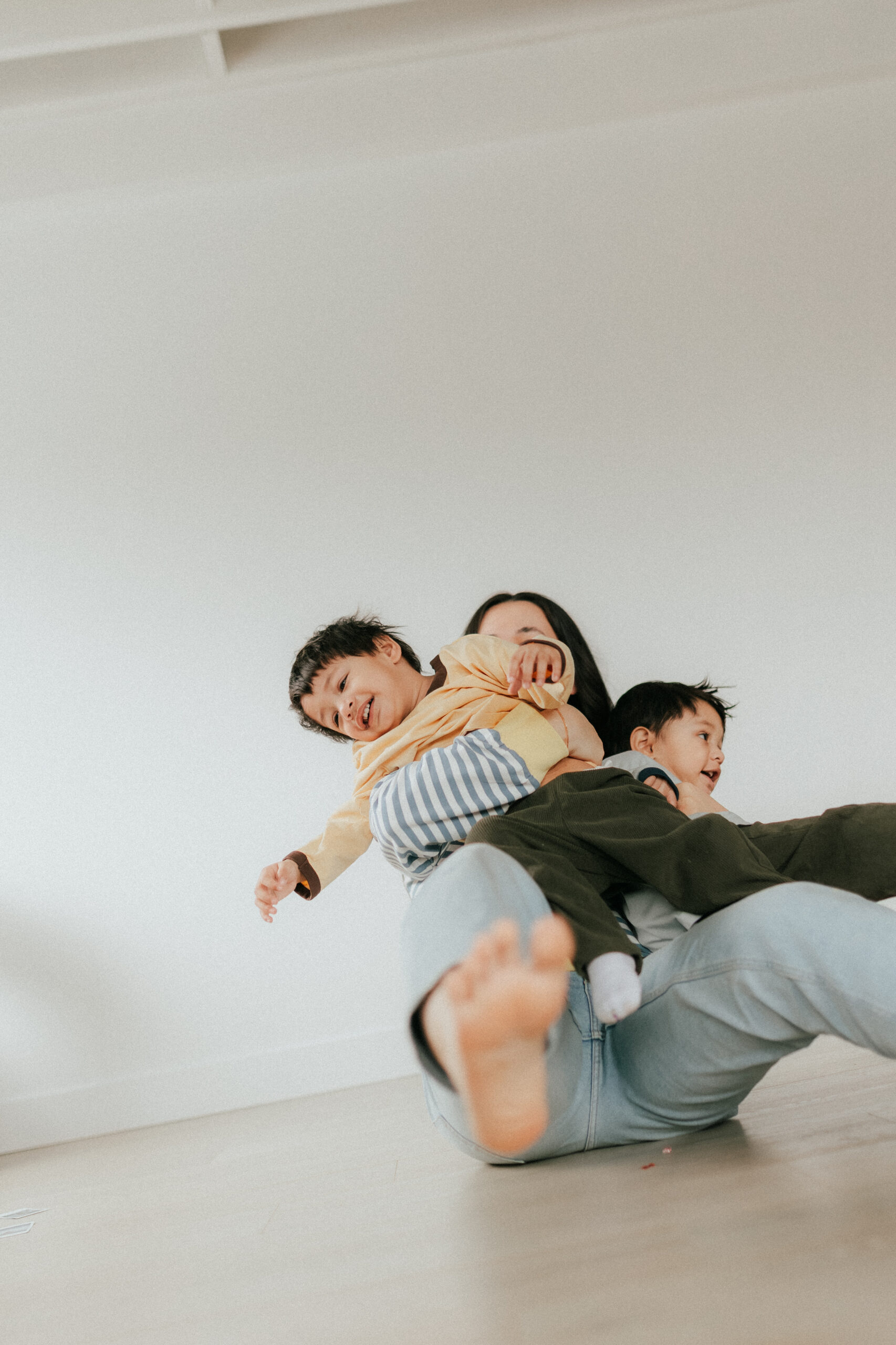 Mom holding her twin toddler sons and sitting on the floor while spinning around in circles to make them laugh for a motherhood session in abbotsford