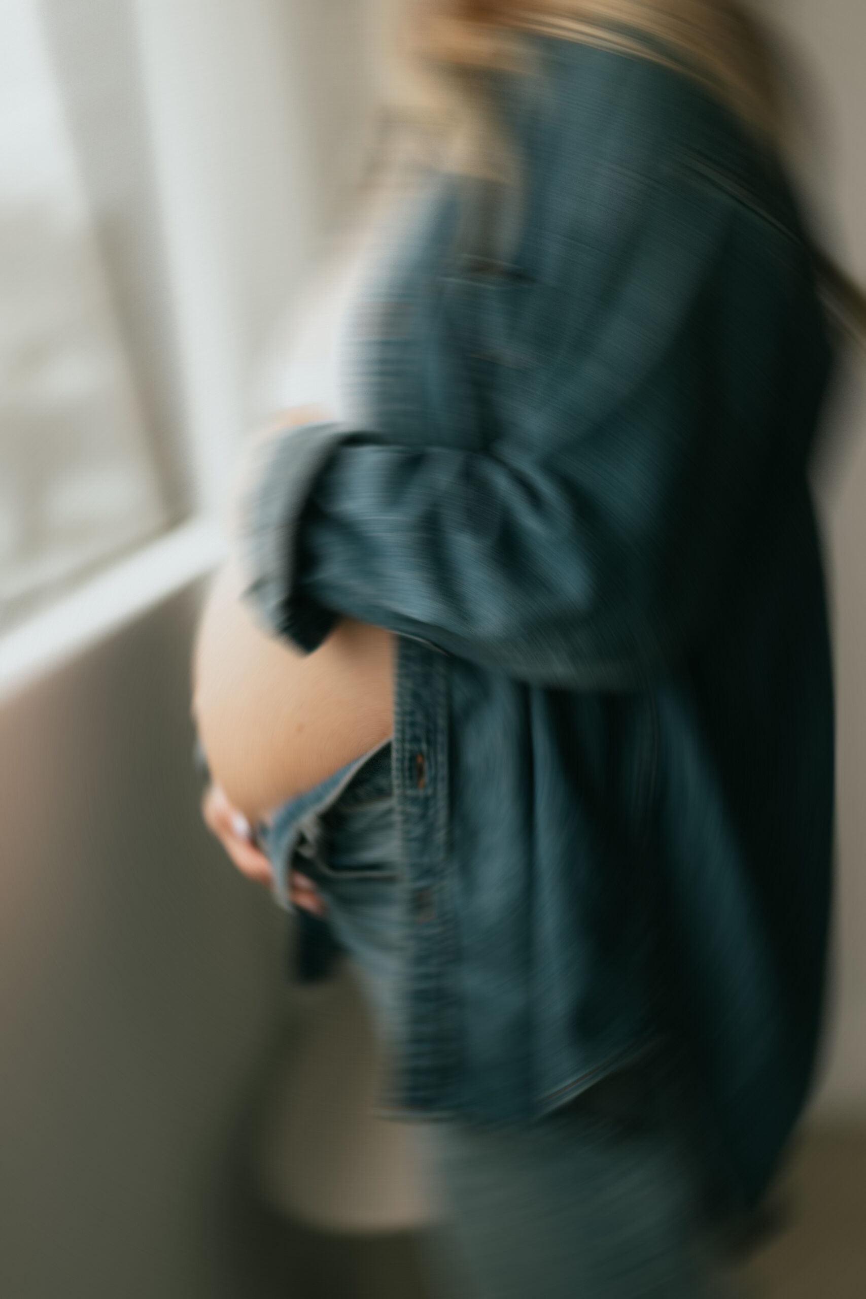 Blurred photo of a pregnant woman holding her baby bump while standing in front of a window taken by a maternity and newborn photographer in abbotsford