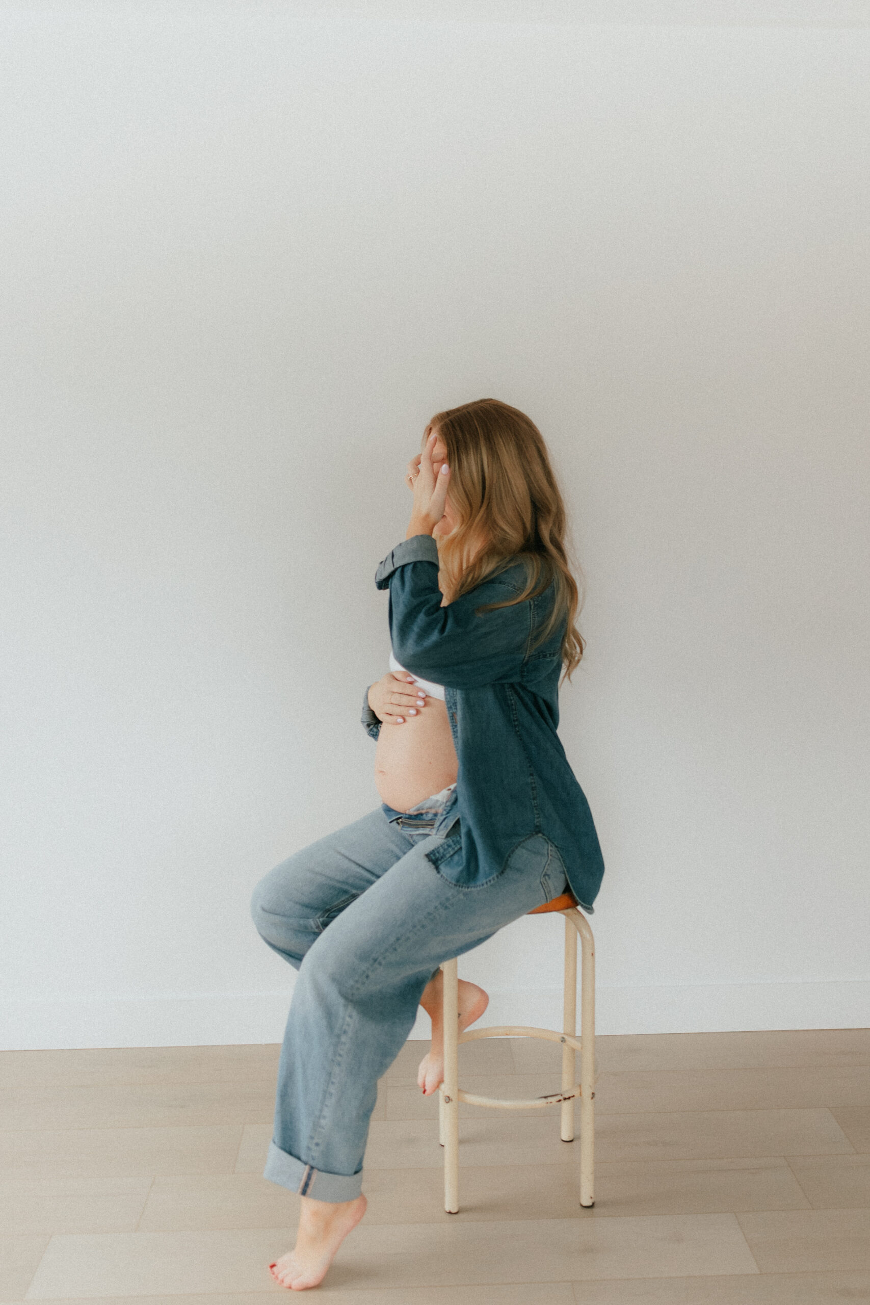 Photo of a pregnant woman sitting on a stool facing towards a window while moving a piece of hair out of her face