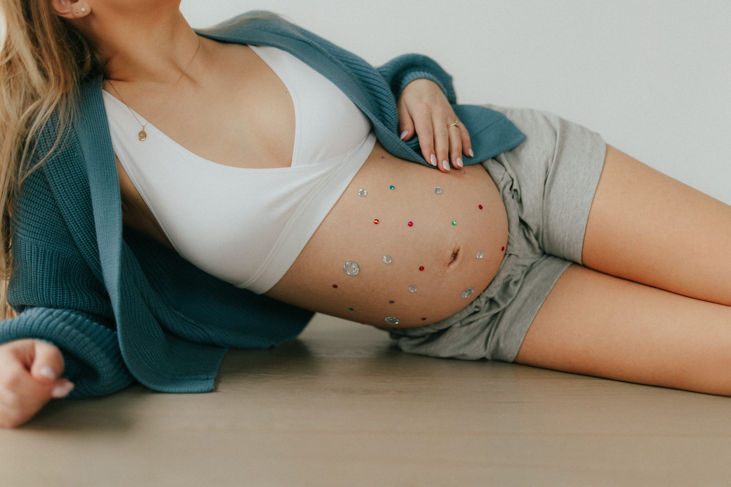 Photo of a pregnant woman laying on her side on the floor with her hand draped on her baby bump that is covered in gemstones
