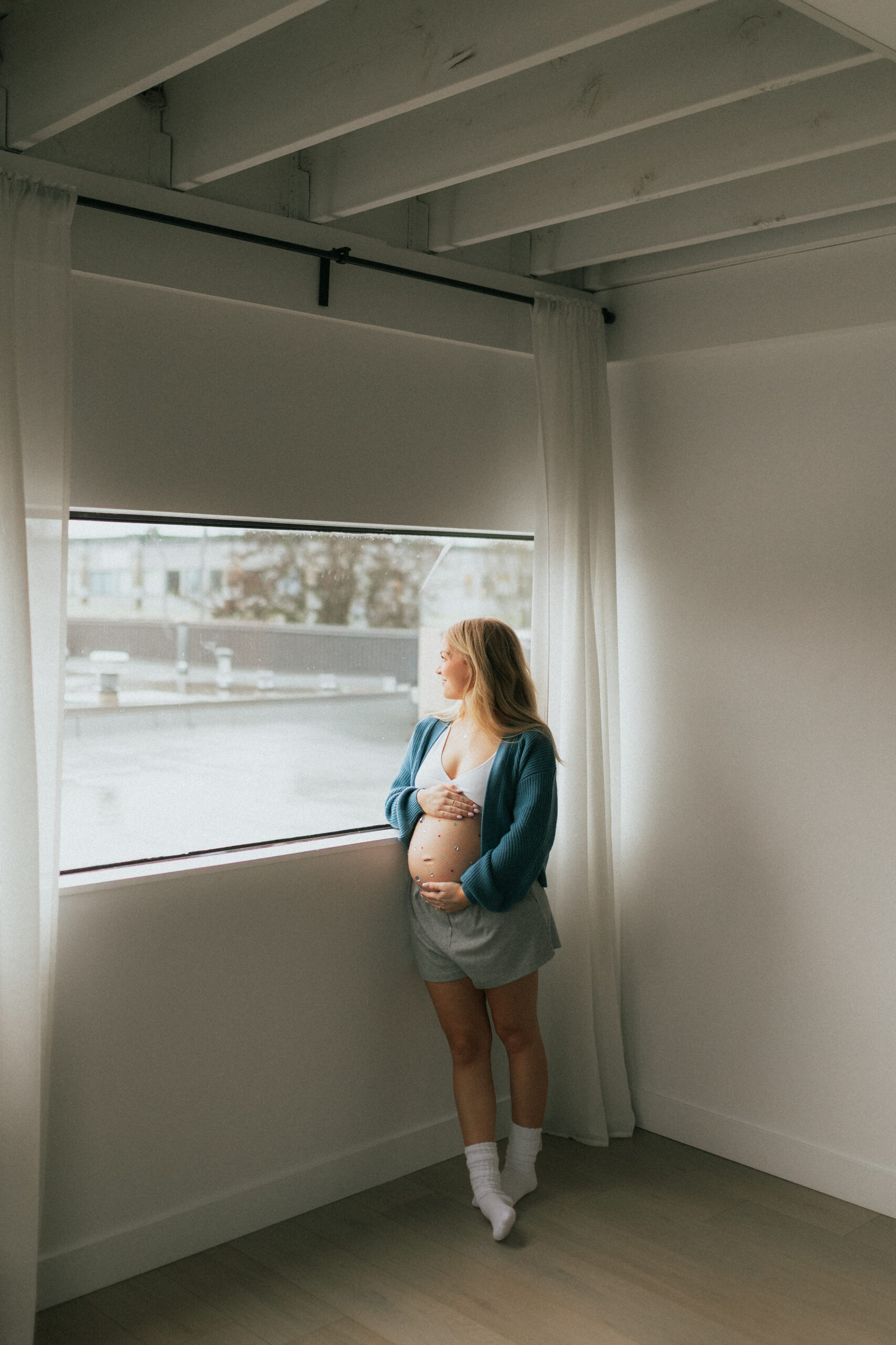 Photo of a pregnant woman holding her baby bump while looking out of the window of a white studio