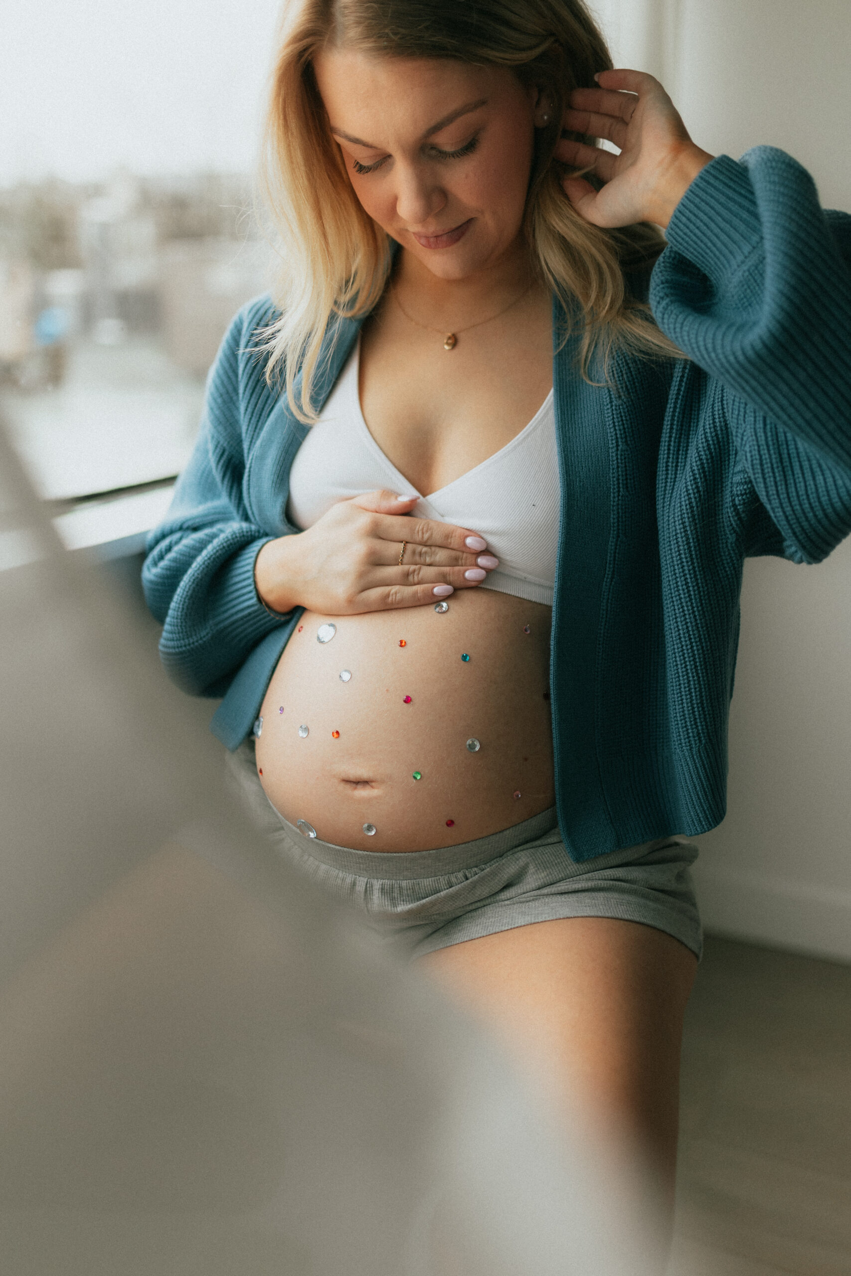 photo of a pregnant woman sitting on a stool looking down at her baby bump that is covered in colourful gem stones